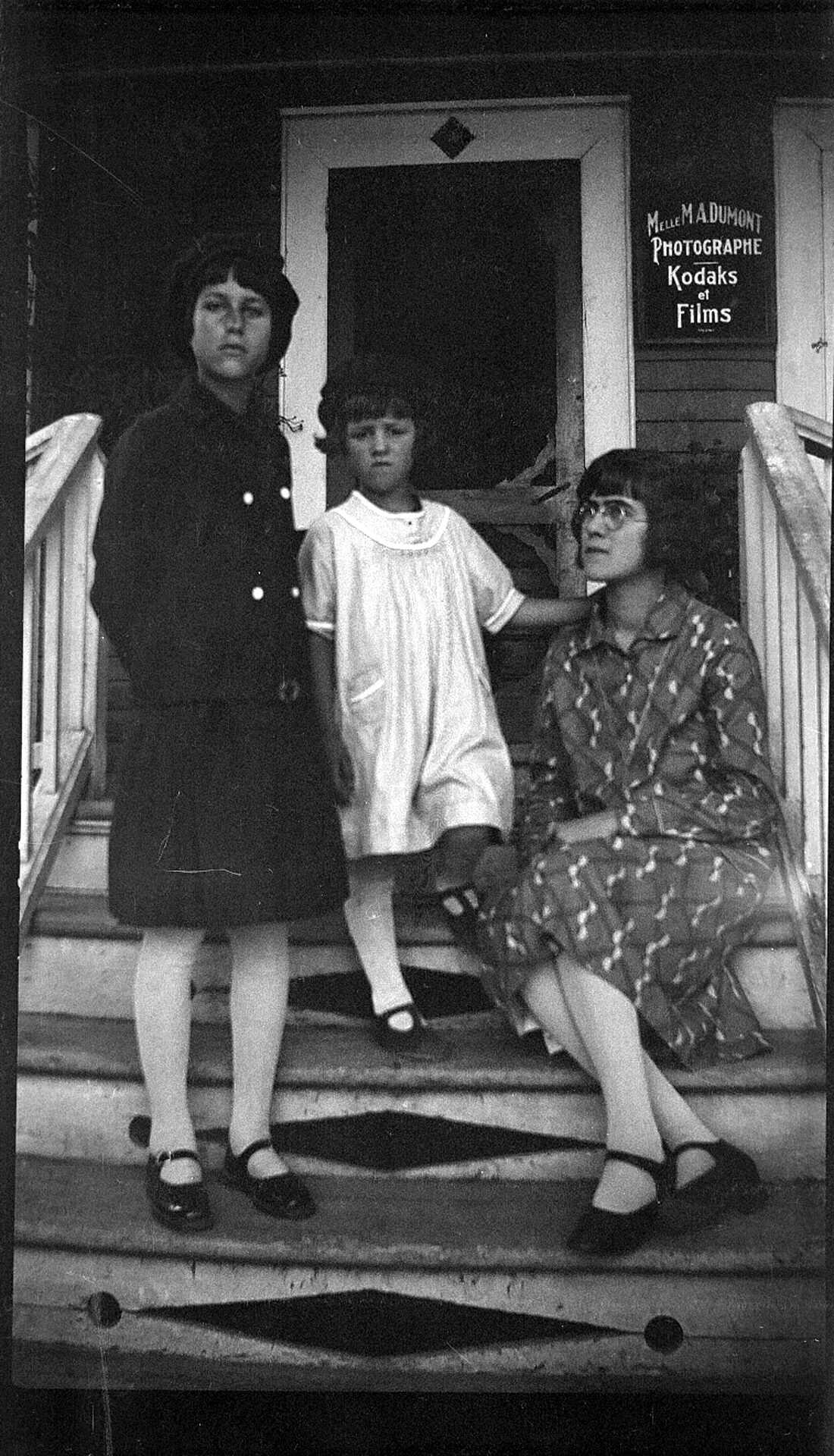 Portrait de trois jeunes filles dont Elizabeth Dumont devant l'entrée du studio Dumont et une affiche indiquant "Melle M. A. Dumont Photographe Kodaks et Films".