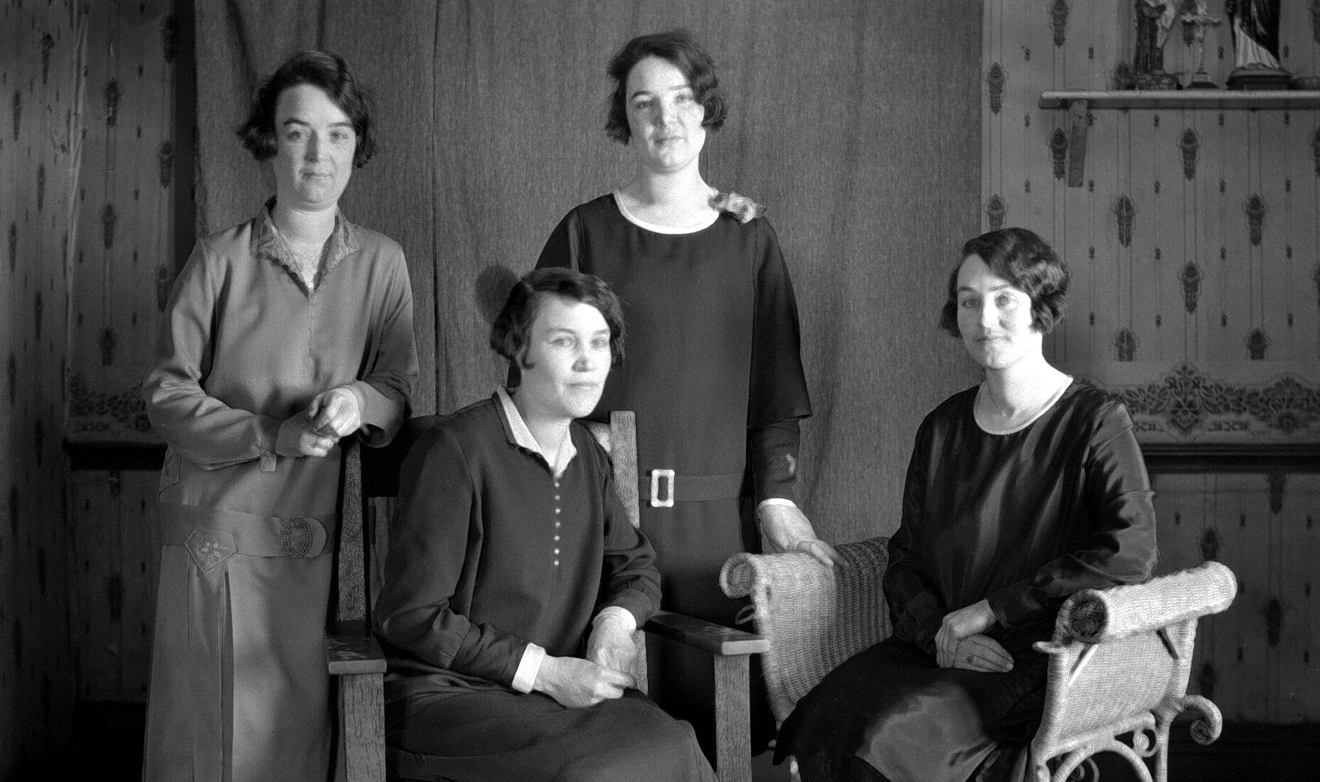 Portrait de quatre jeunes femmes dont deux sont assises dans des fauteuils en bois et en osier dans un décor domestique.