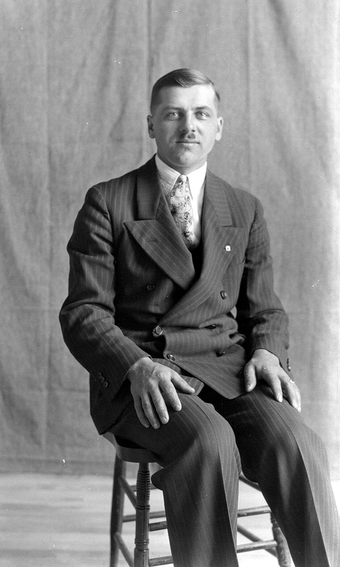 Portrait d'un homme âgé d'environ 30 ans assis sur un tabouret devant un grand drap gris.
