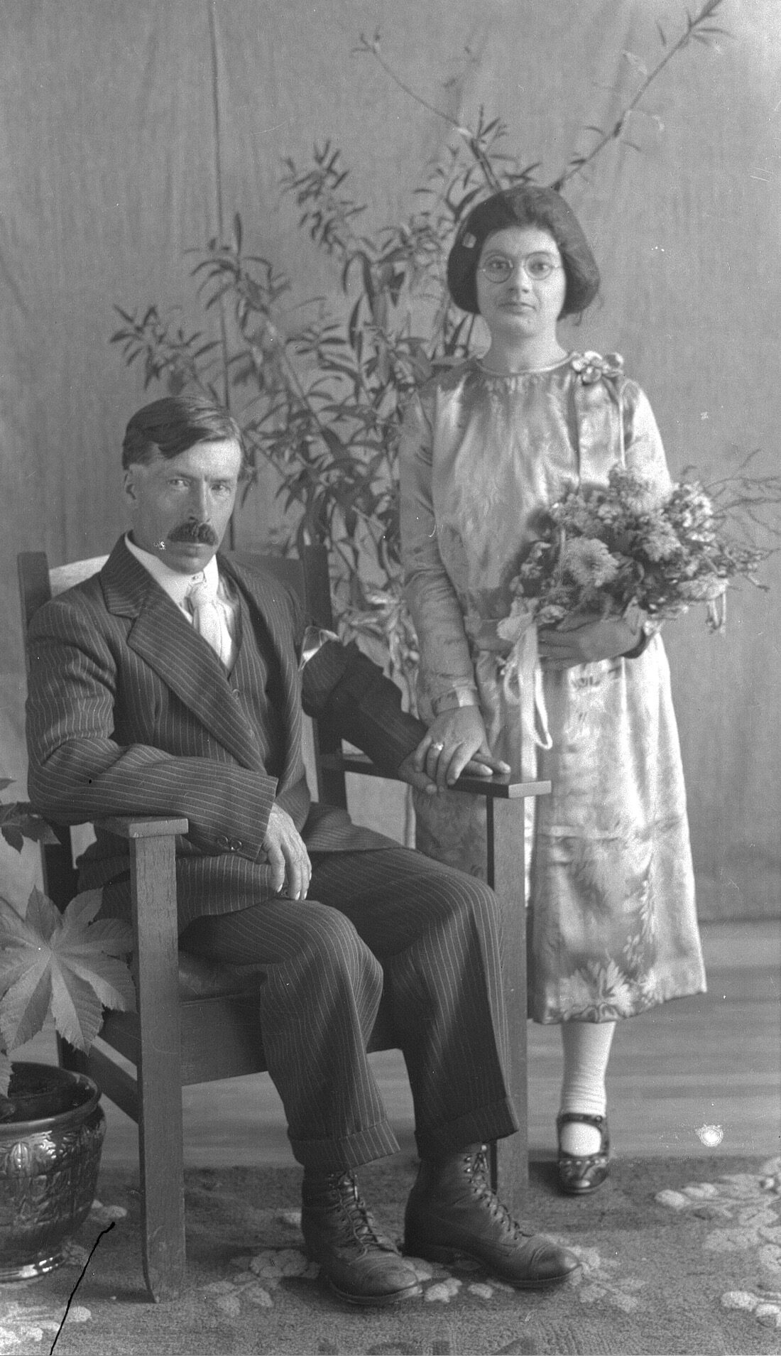 Portrait d'un jeune couple dans un décor comprenant une grande plante en arrière-plan et dont la femme, debout, tient un bouquet de fleur et dont l'homme est assis dans un fauteuil en bois.