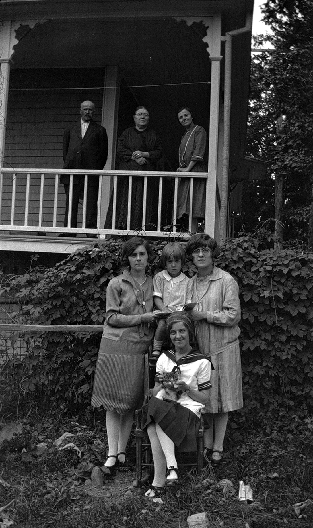 Marie-Alice Dumont, ses parents, une soeur et d'autres jeunes filles prenant la pose sur la galerie et sur le terrain de la maison des Dumont.