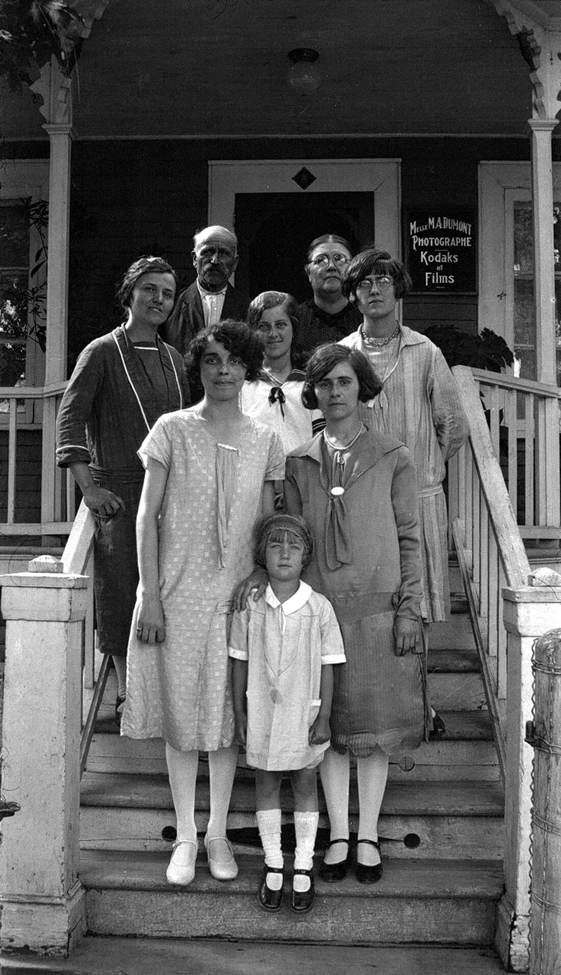 Huit personnes d'âges divers dont Marie-Alice Dumont debout dans l'escalier d'un perron, un panneau en arrière-plan indique "Mlle M. A. Dumont Photographe Kodaks et Films".