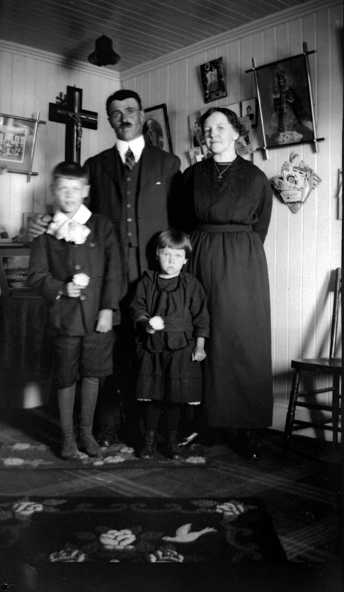 François Dumont et Mary April avec leurs deux enfants tous vêtus élégamment dans un environnement domestique comprenant des photographies et des symboles religieux accrochés aux murs.