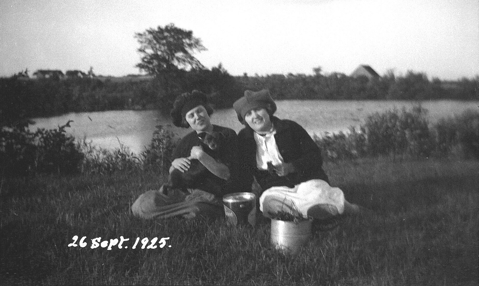 Deux jeunes femmes dont l'une tient un petit chien dans ses bras assises dans le gazon sur le bord d'une rivière.
