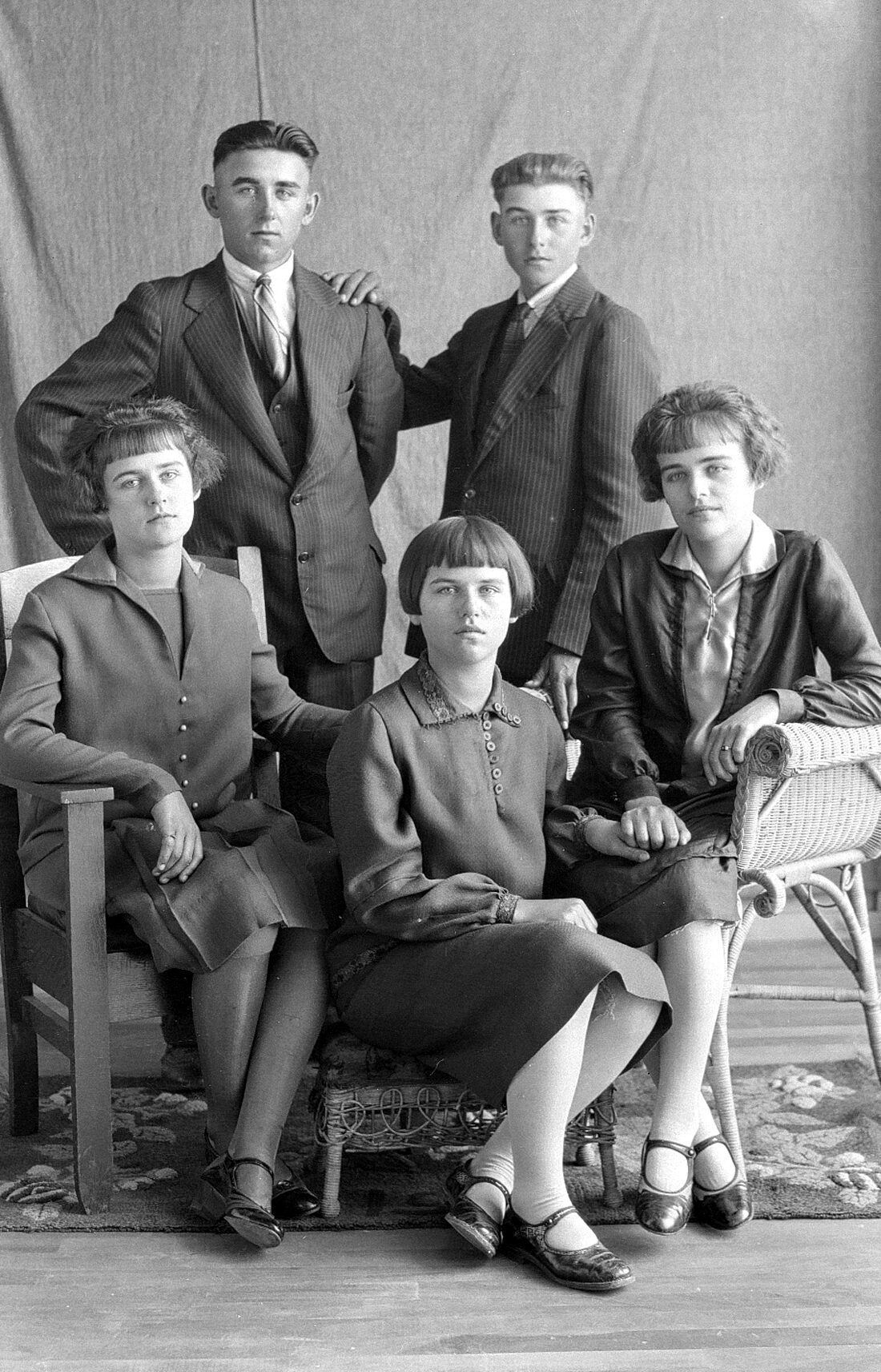 Portrait de studio de trois jeunes femmes assises sur des fauteuils et de deux jeunes hommes debout derrière elles dans un décor dépouillé.
