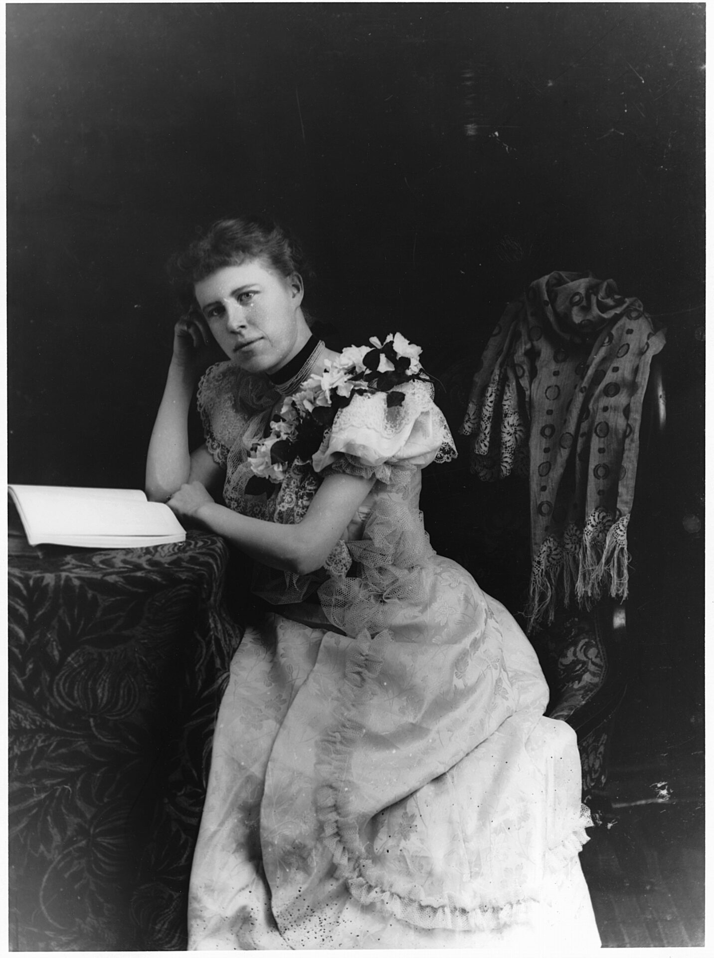 Portrait d'une femme élégante assise devant un livre ouvert sur une table et dont la tête repose sur la main de manière nonchalante.