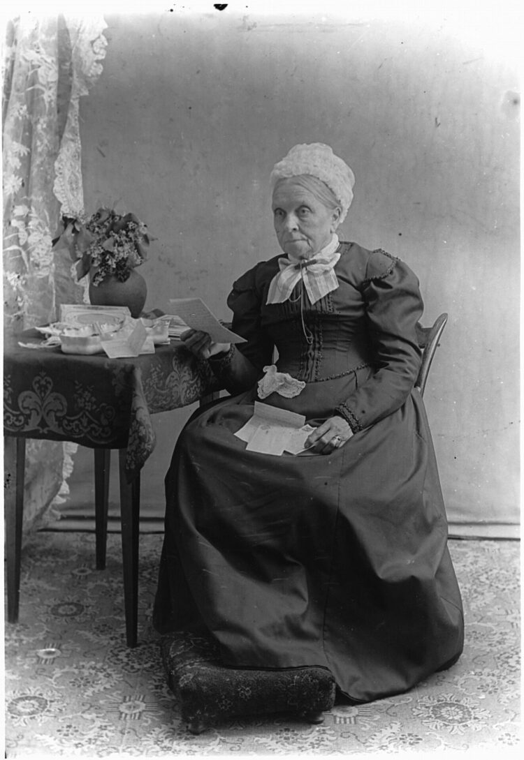 Portrait d'une vieille dame vêtue à la mode victorienne attablée et lisant une lettre.