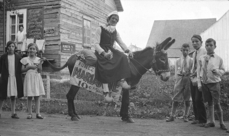 Un vendeur de tabac Marianne costumé en paysanne et assis sur son âne, posant devant un magasin général et flanqué de trois jeunes garçons et deux jeunes filles.
