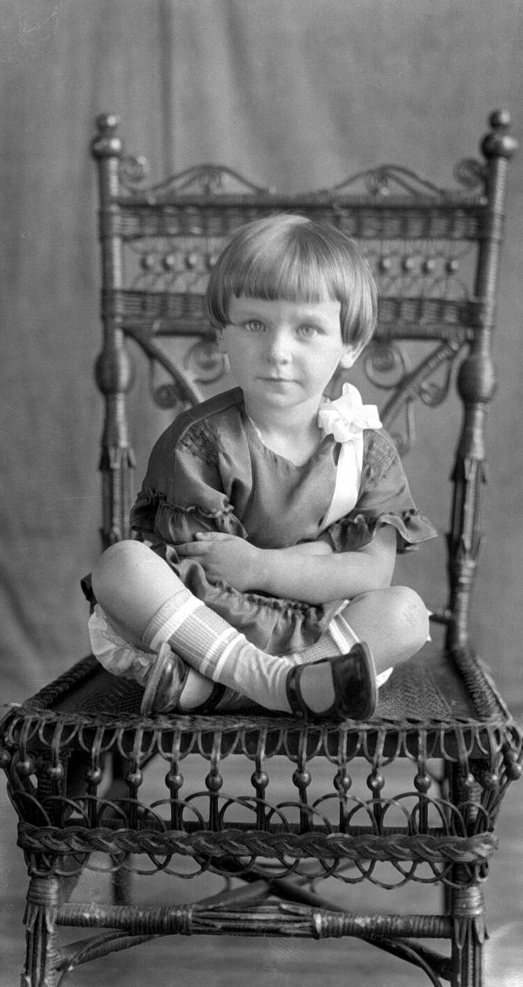 Studio portrait of a little girl sitting cross-legged with crossed arms on a chair.
