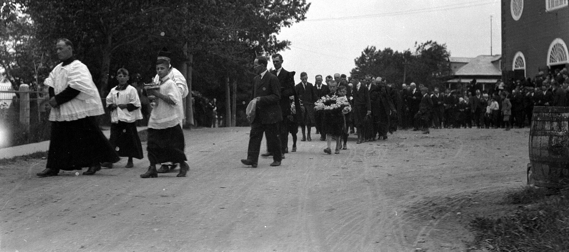 Une foule vêtue de noir sortant d'une église et suivant, en file, un prêtre se dirigeant vers un cimetière.