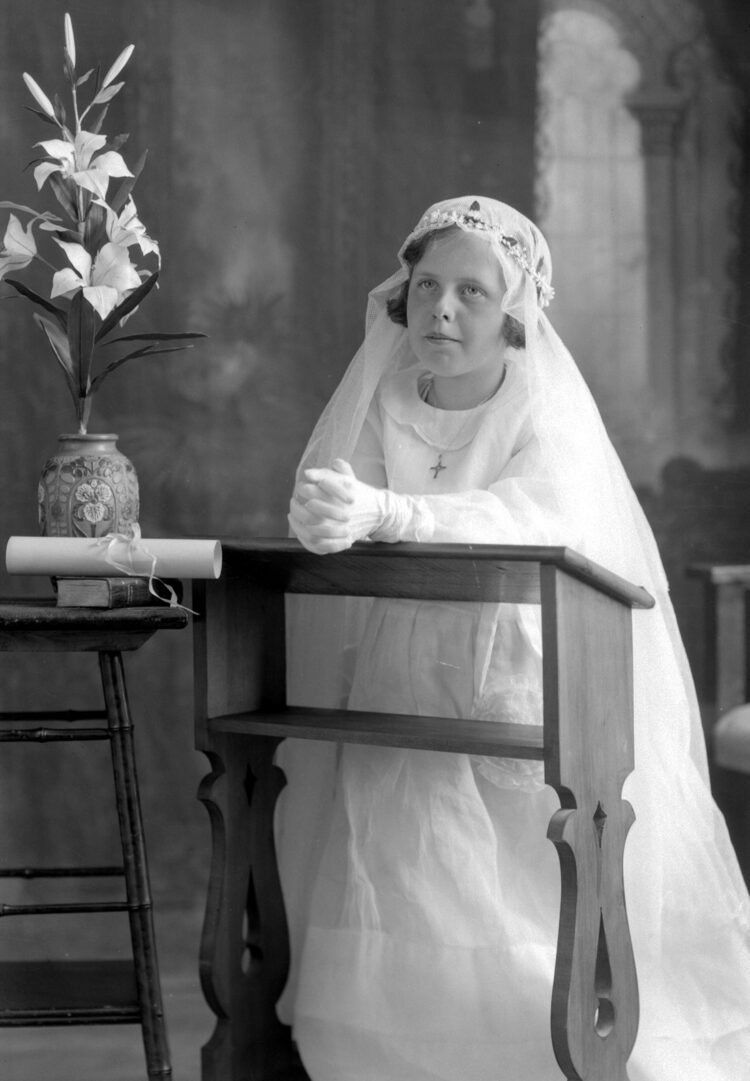 Jeune fille en studio agenouillée à un prie-Dieu vêtue d'une robe, de gants et d'un voile tous de couleur blanche et flanquée d'une fleur de lys, d'un diplôme et d'un livre sur un tabouret.