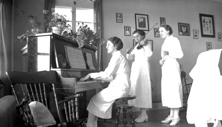 Trois femmes en uniforme de garde-malade posant à l'intérieur, l'une joue du piano, l'autre du violon et l'autre chante.