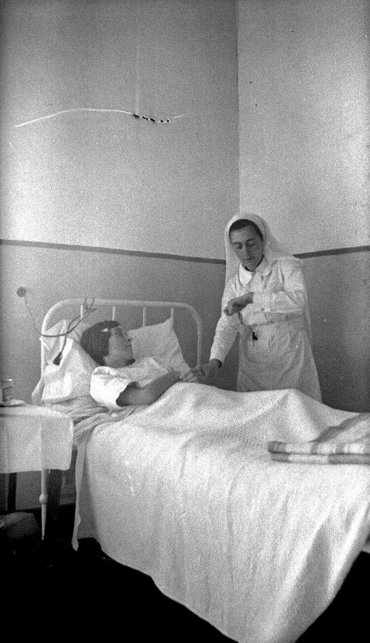 Une femme alitée et malade, une autre en uniforme de garde-malade debout à ses côtés et prenant son pouls.