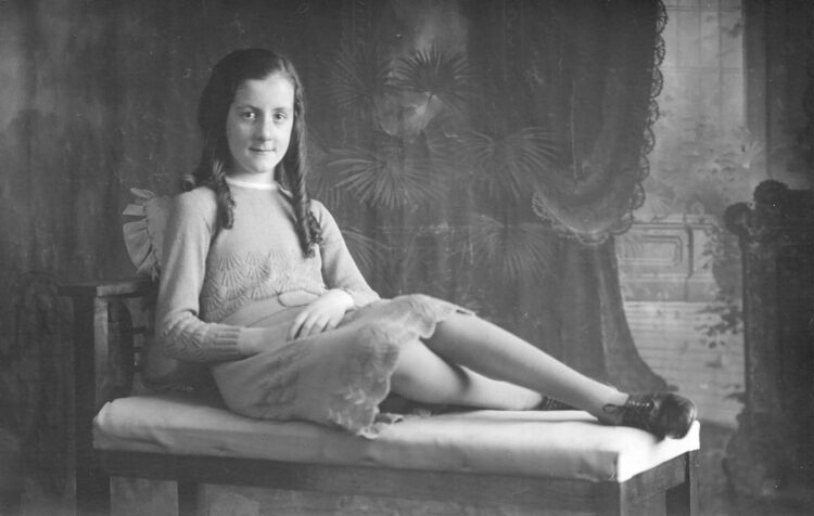 Studio portrait of a young girl sitting on a bench, her legs extended and crossed in front of her.