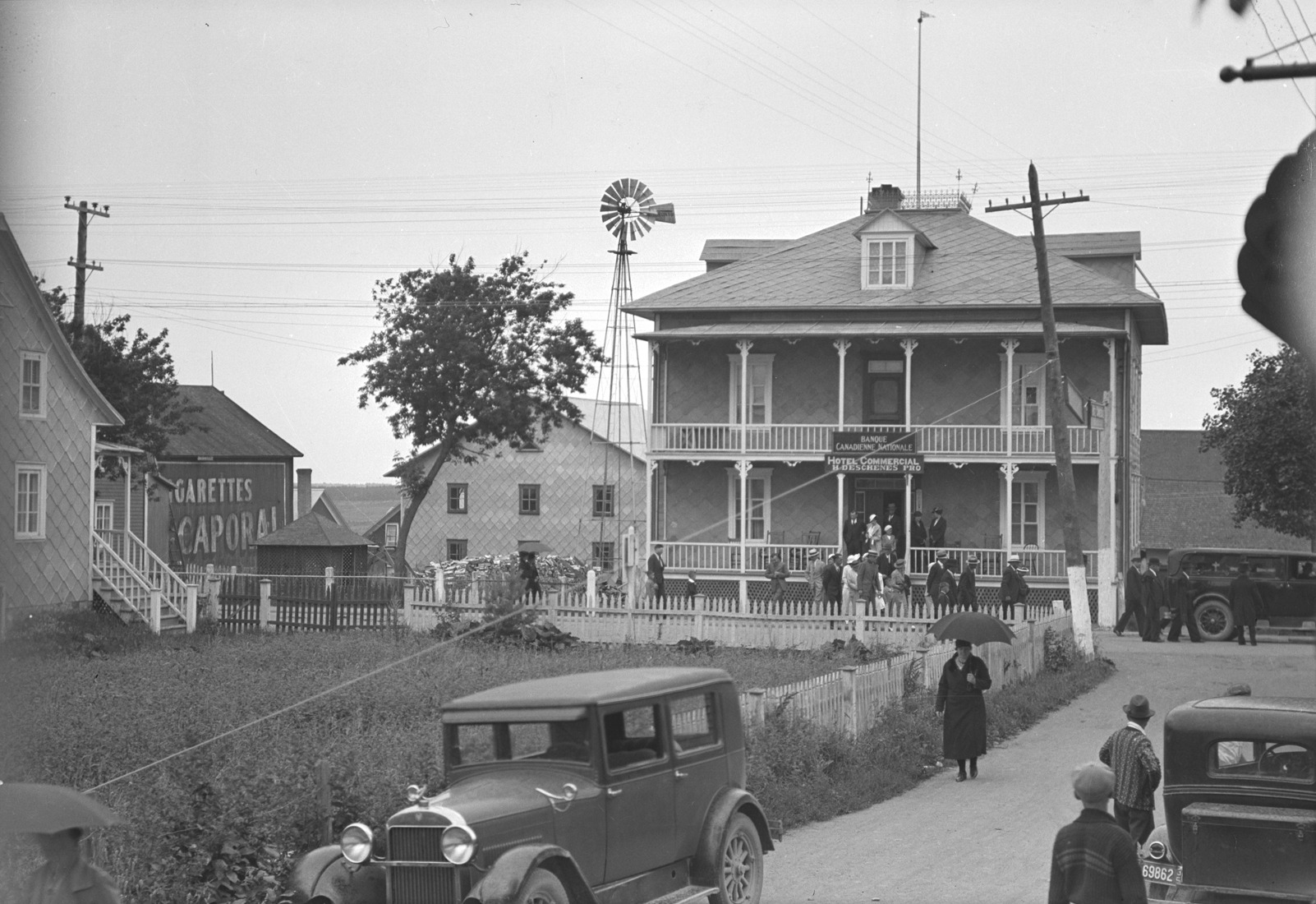Scène du village de Saint-Alexandre incluant des piétons, des automobiles et des bâtiments dont celui abritant la "Banque Canadienne nationale" et l'"Hôtel Commercial H. Deschênes Pro".