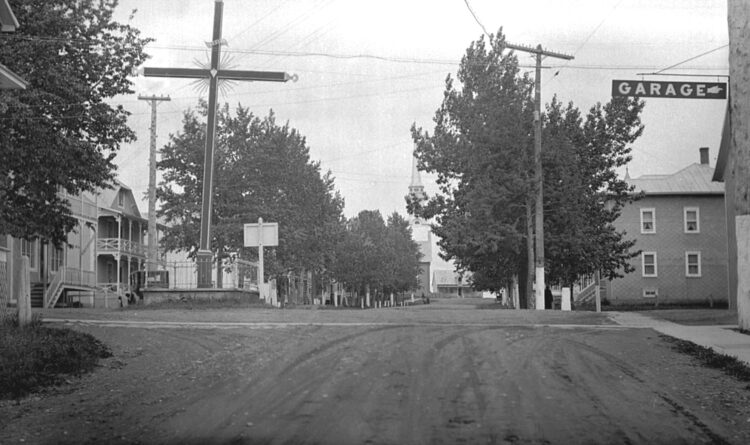 Croisement de deux routes au centre d'un village dont plusieurs des infrastructures sont visibles telles que des maisons, trottoirs, poteaux et fils électriques.