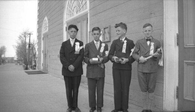Quatre garçons en habits de confirmation, diplômes en main, sur le perron de l'église.