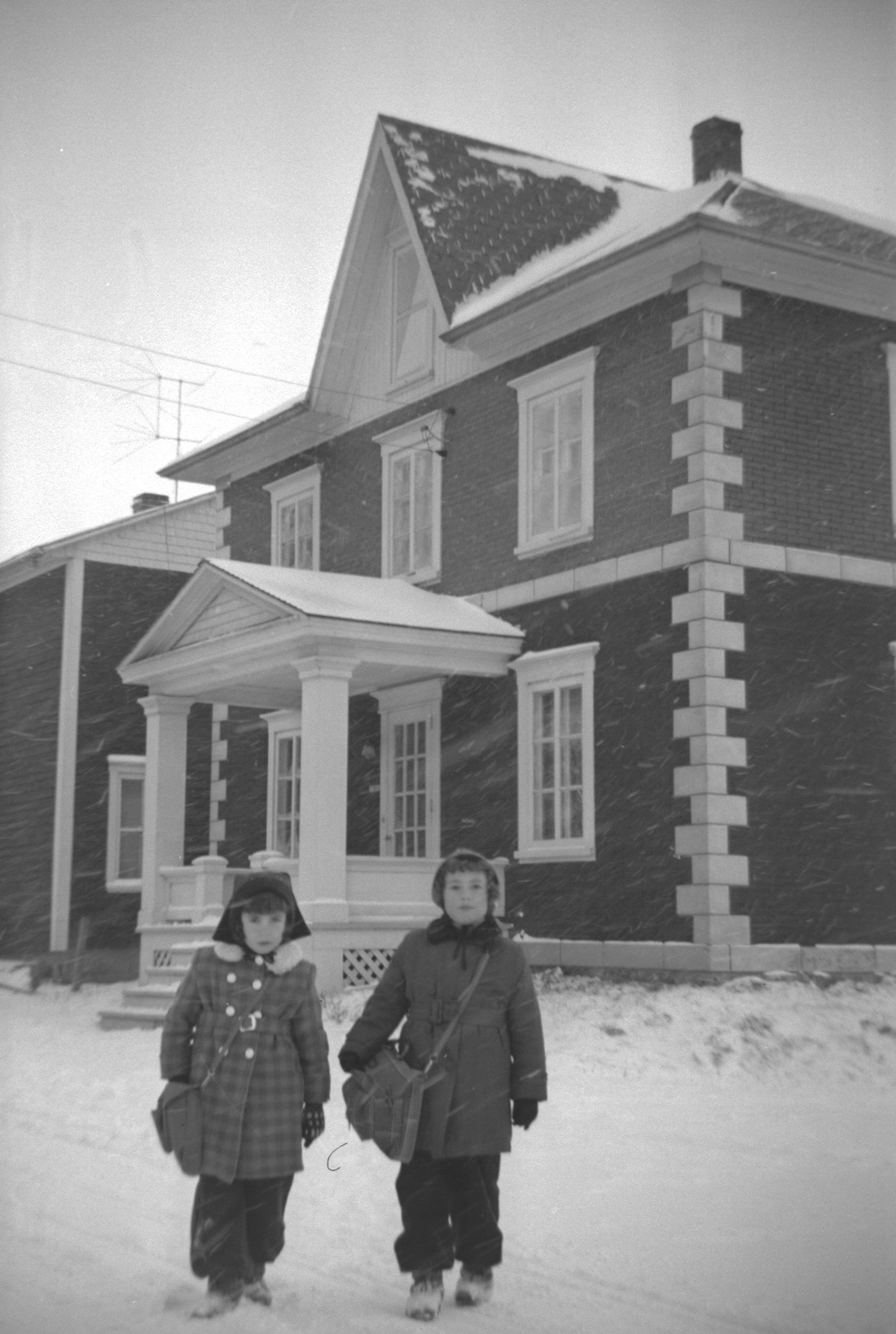 Deux jeunes filles s'apprêtant à aller à l'école posant, en hiver, des flocons volant au vent, devant une maison.