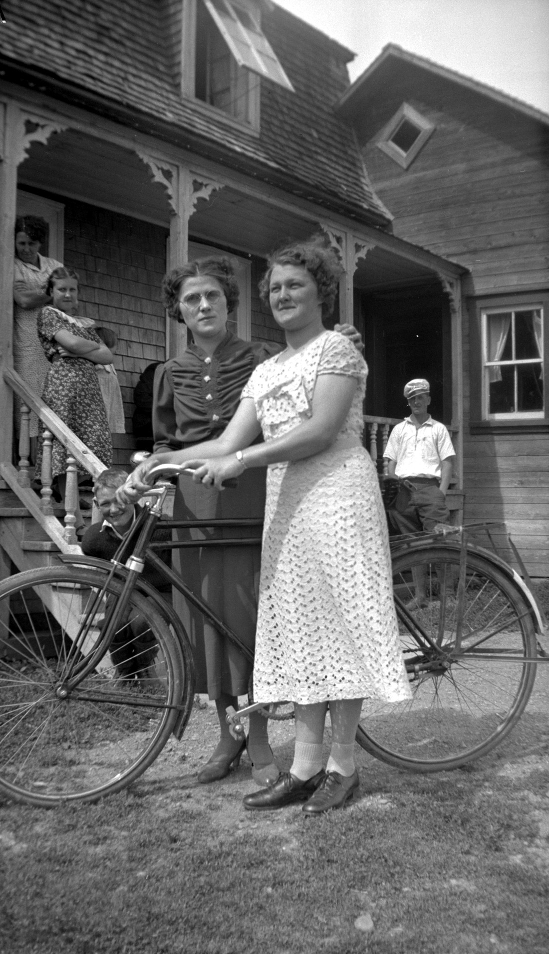 Deux femmes debout de chaque côté d'une bicyclette avec, en arrière-plan, une résidence et quatre autres personnes les observant.