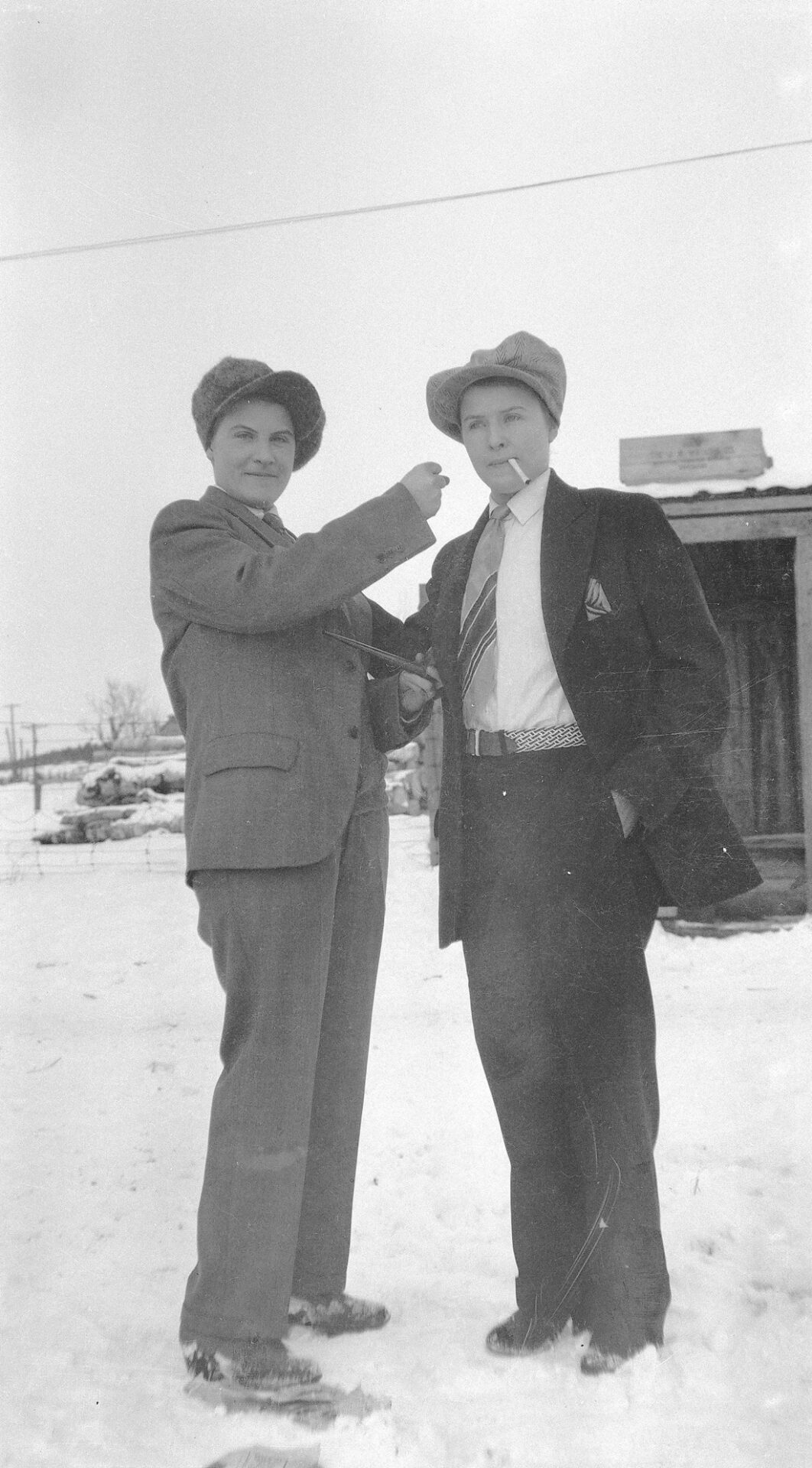Deux jeunes femmes posant en hiver, à l'extérieur, en costume pour homme, l'une pipe à la main et faisant mine de frapper au visage l'autre qui tient une cigarette entre ses lèvres.