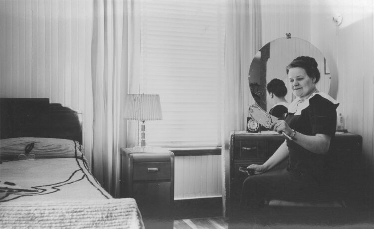 Femme assise à une coiffeuse dans une chambre à coucher et se regardant dans un petit miroir qu'elle tient dans sa main droite.