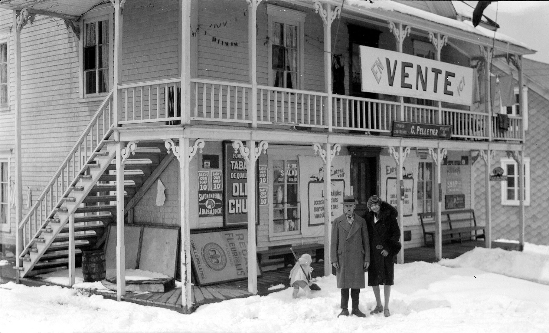 Un couple posant, en hiver, devant un magasin général et avec, derrière eux, un enfant d'environ deux ans pelletant de la neige.