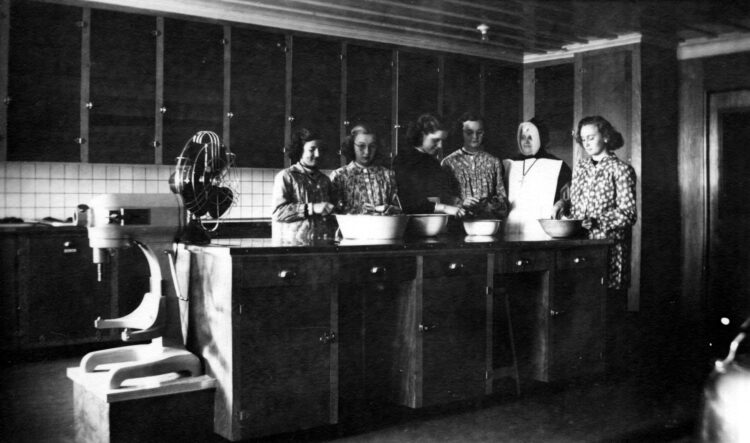 Cinq couventines et leur religieuse enseignante dans une cuisine, à la préparation de légumes ou de fruits.