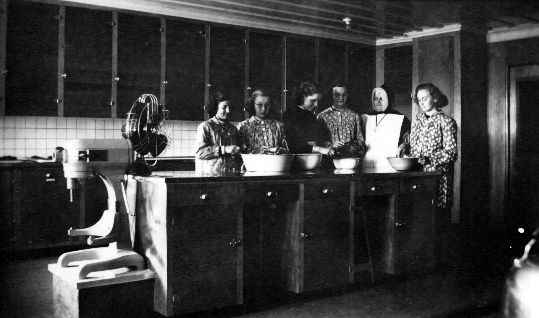 Cinq couventines et leur religieuse enseignante dans une cuisine, à la préparation de légumes ou de fruits.