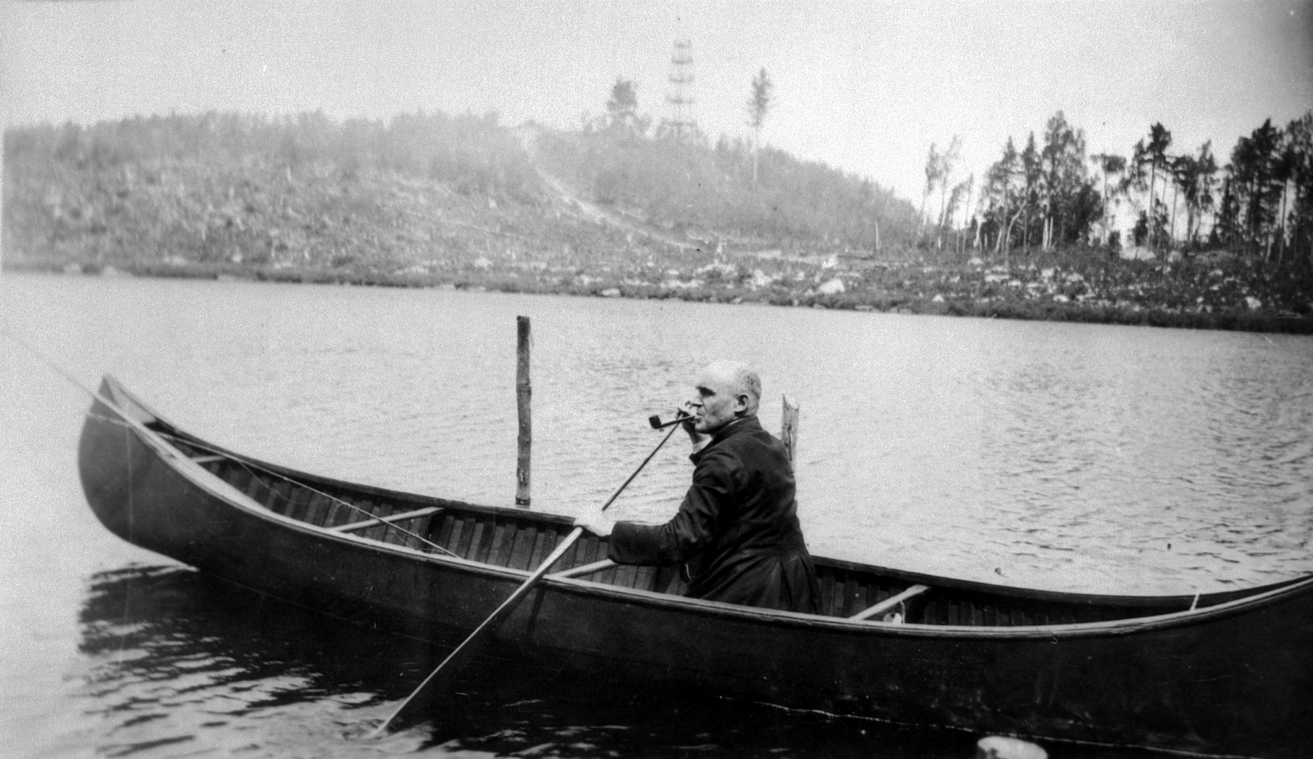 Un religieux agenouillé dans un canot, pagaie à la main et pipe au bec, sur un lac dont les bords sont fraîchement défrichés.