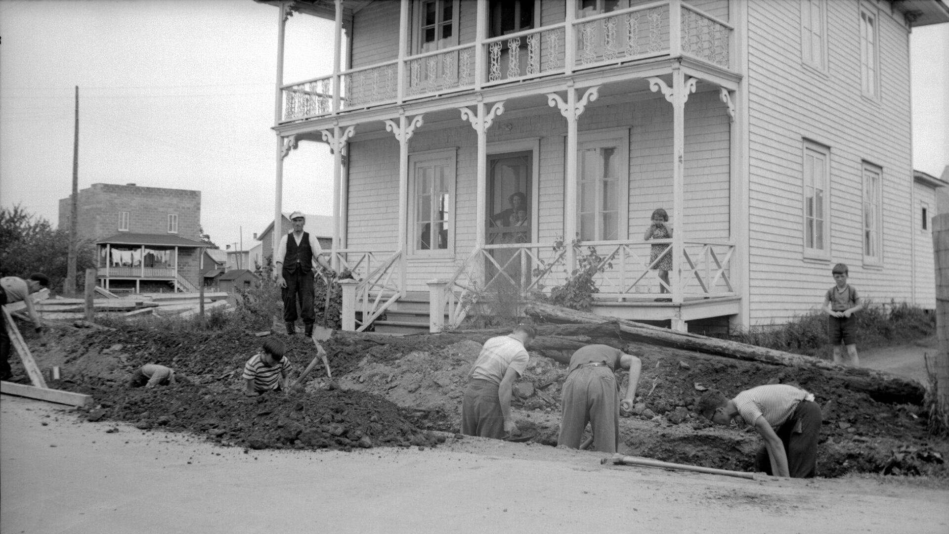Sept jeunes hommes creusant une tranchant devant une grande résidence et sous le regard d'une femme et trois enfants.