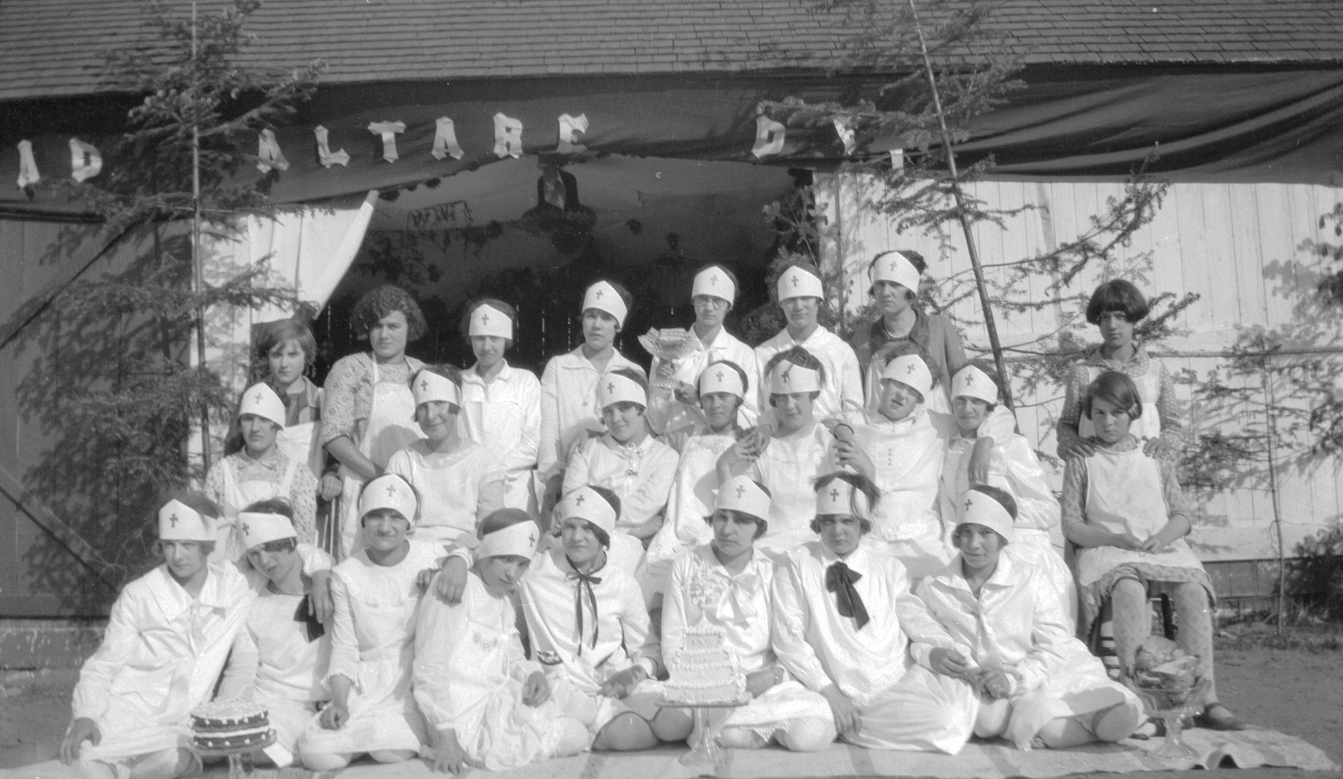 24 jeunes filles en uniforme blanc de scout devant un bâtiment sur lequel une banderole indiquant "Ad Altare Dei" est installée.