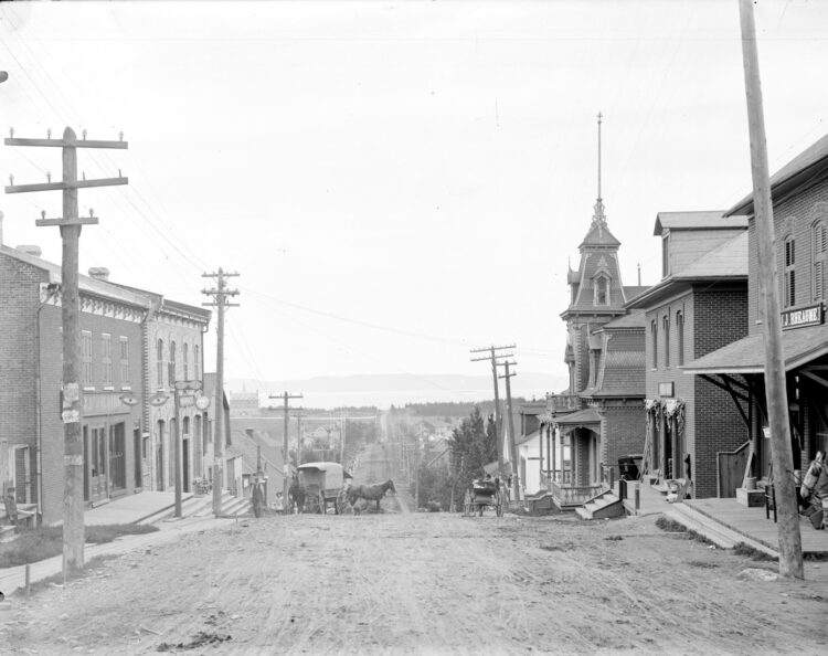Vue portant au loin d'une rue de Rivière-du-Loup bordée d'édifices commerciaux et dans laquelle des calèches sont tirées par des chevaux.