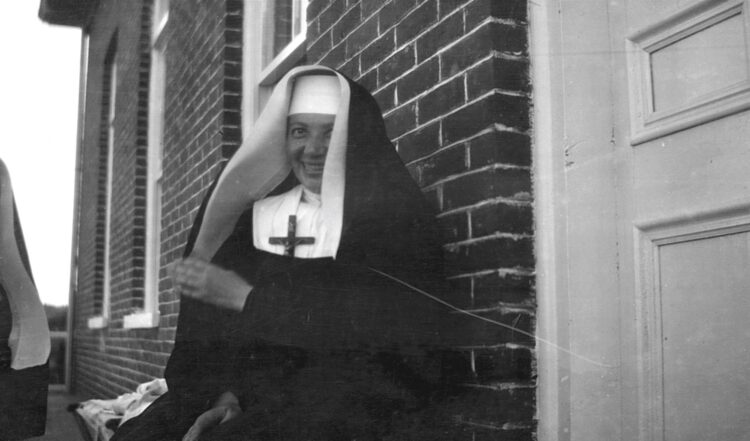 Portrait d'une religieuse souriant à la caméra sur la galerie d'un édifice en briques.