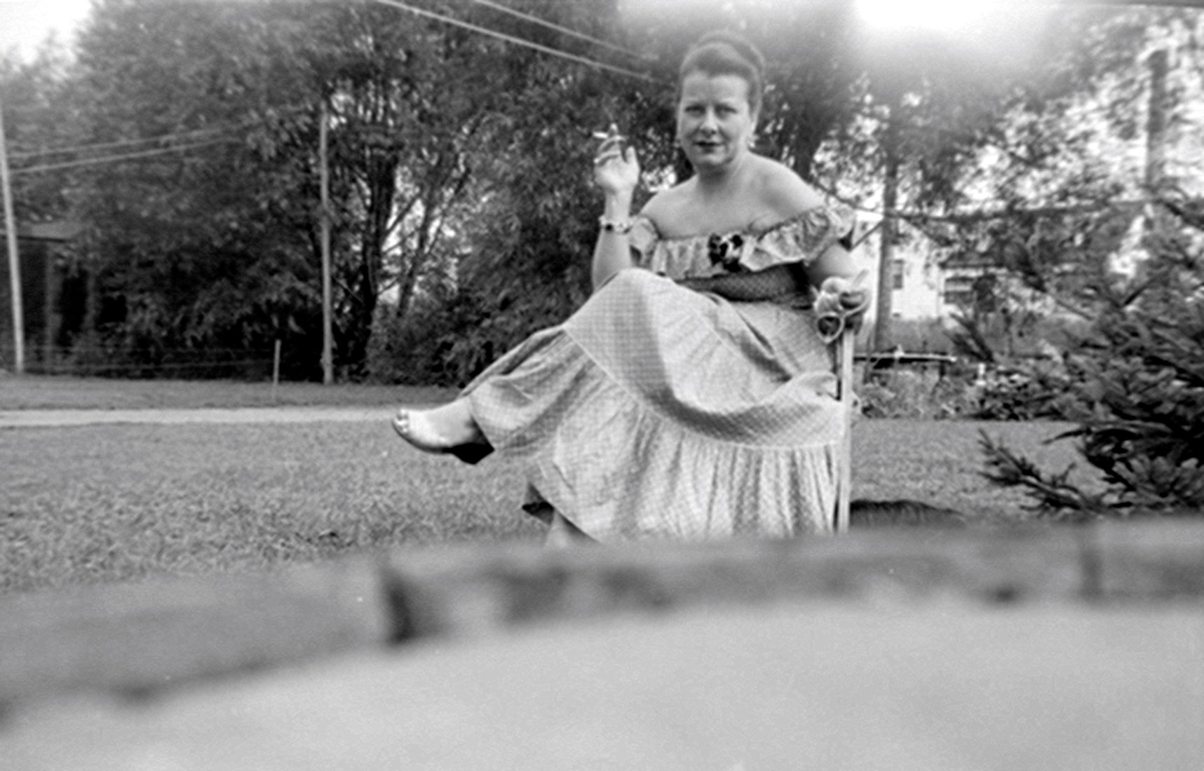 Portrait en extérieur d'une femme en robe échancrée assise sur une chaise jambes croisées et fumant une cigarette.