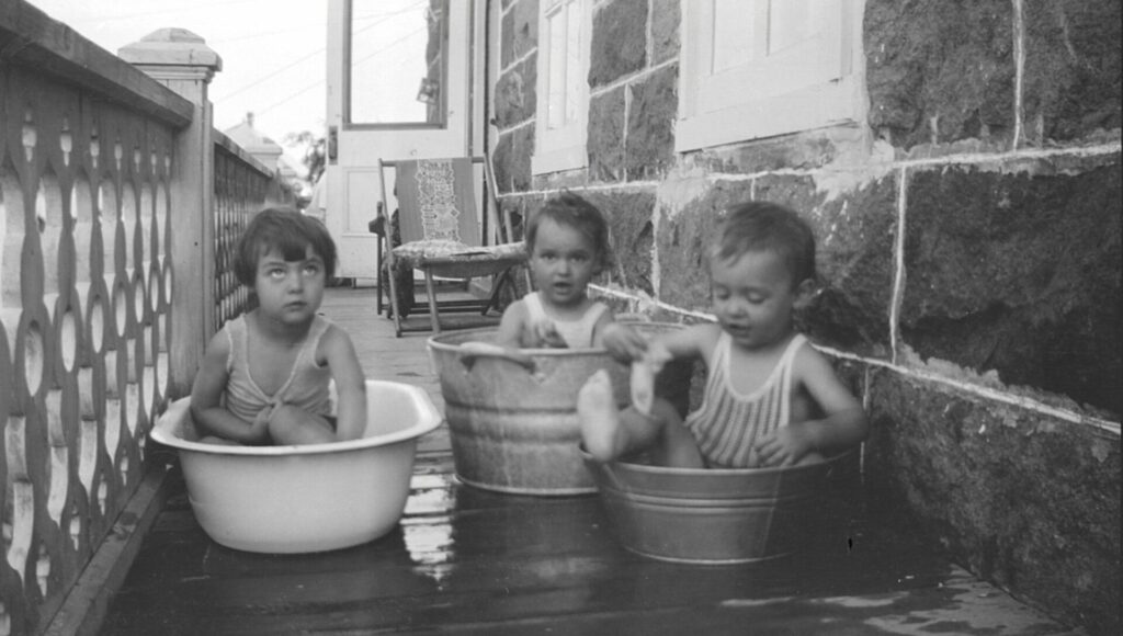 Trois jeunes enfants installés dans des bassines sur la galerie d'une maison faite de grosses pierres rectangulaires.