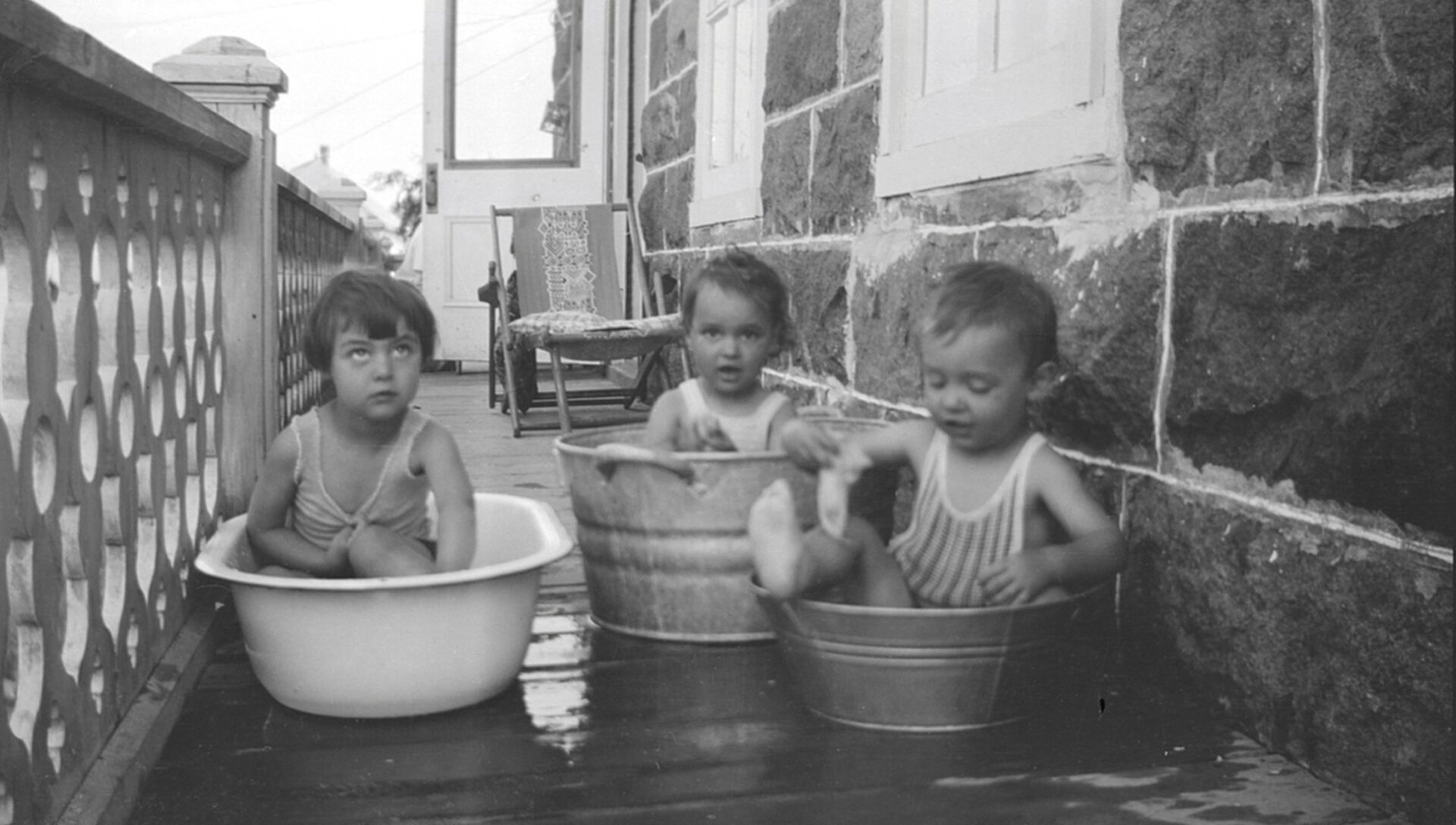 Trois jeunes enfants installés dans des bassines sur la galerie d'une maison faite de grosses pierres rectangulaires.