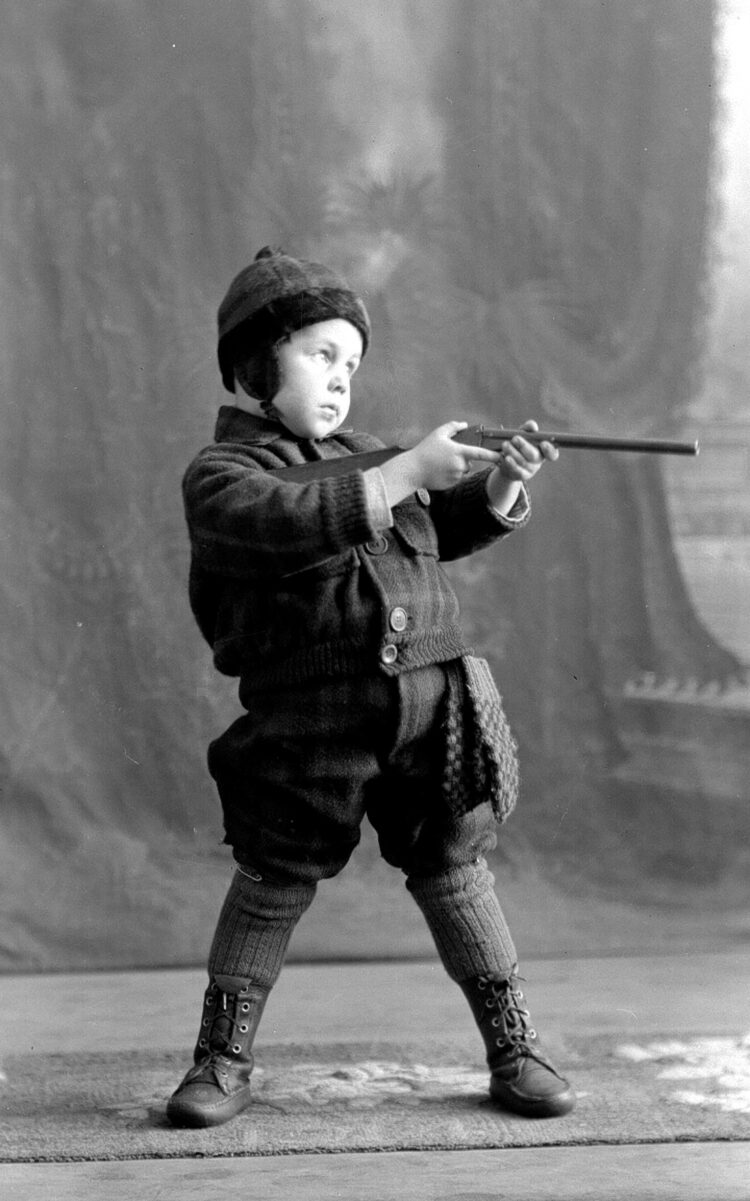 Portrait studio d'un garçonnet vêtu en chasseur et maniant une carabine jouet.