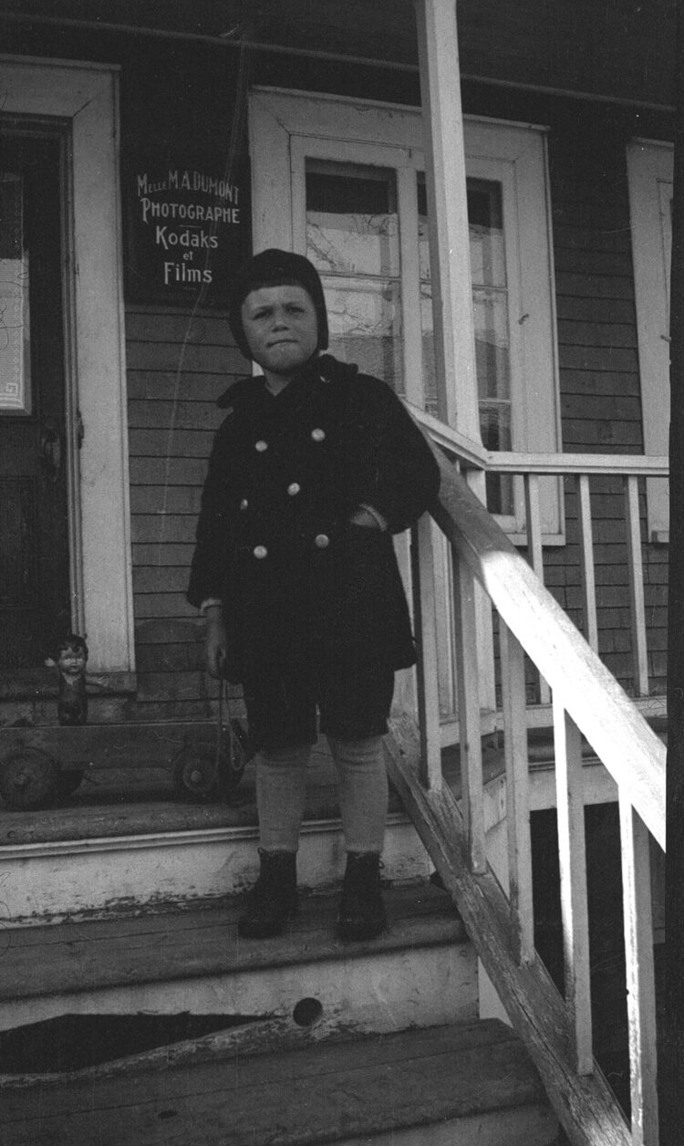 Un jeune garçon et son chariot jouet sur le perron du Studio Dumont, une affiche en arrière-plan indique "Melle M. A. Dumont Photographe Kodaks et Films".