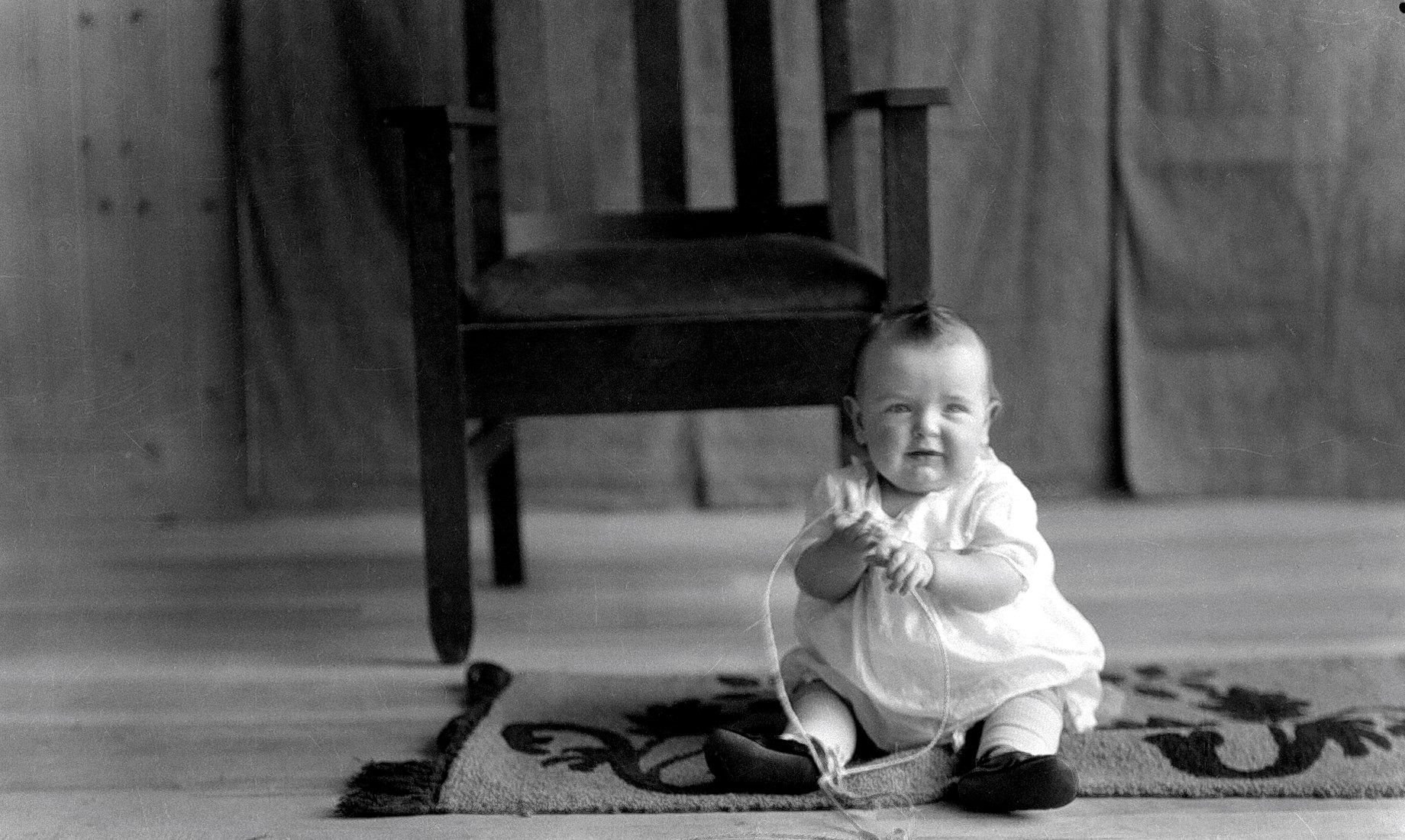 Portrait d'un bébé vêtu d'une robe blanche assis sur un tapis jouant avec ficelle et souriant à la caméra.