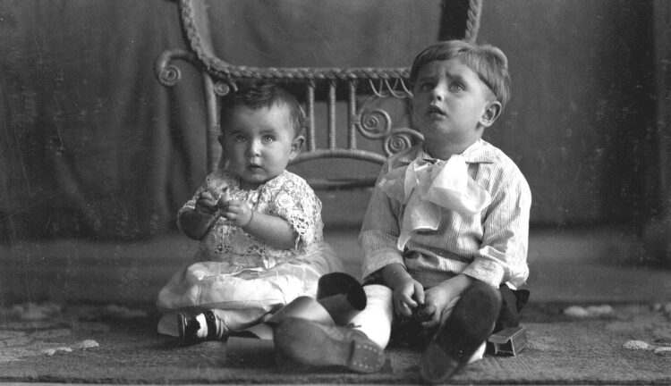 Portrait d'un bébé et d'un garçonnet au regard perplexe assis sur un tapis avec une chaise en osier et un drap gris en arrière-plan.