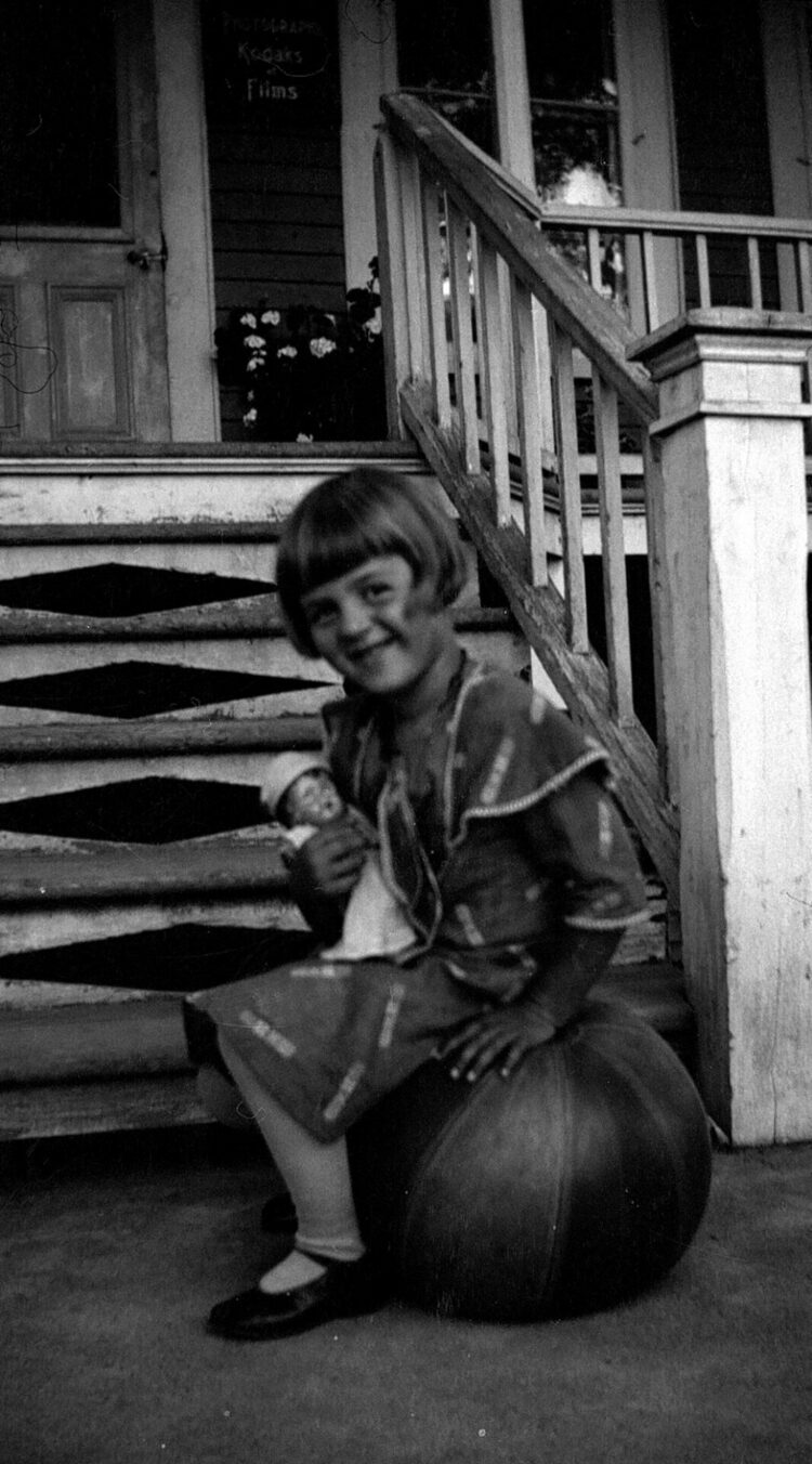 Fillette assise sur un coussin et tenant une poupée posant devant le perron du Studio Dumont".