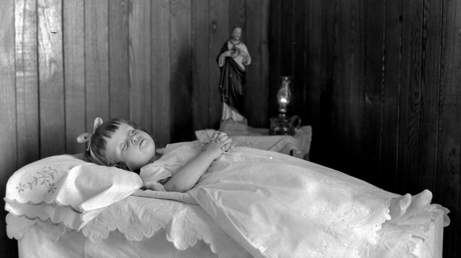 Portrait d'un fillette décédée que l'on a couchée les mains croisées sur le ventre, revêtue d'une robe blanche et placée devant une statuette du Christ.