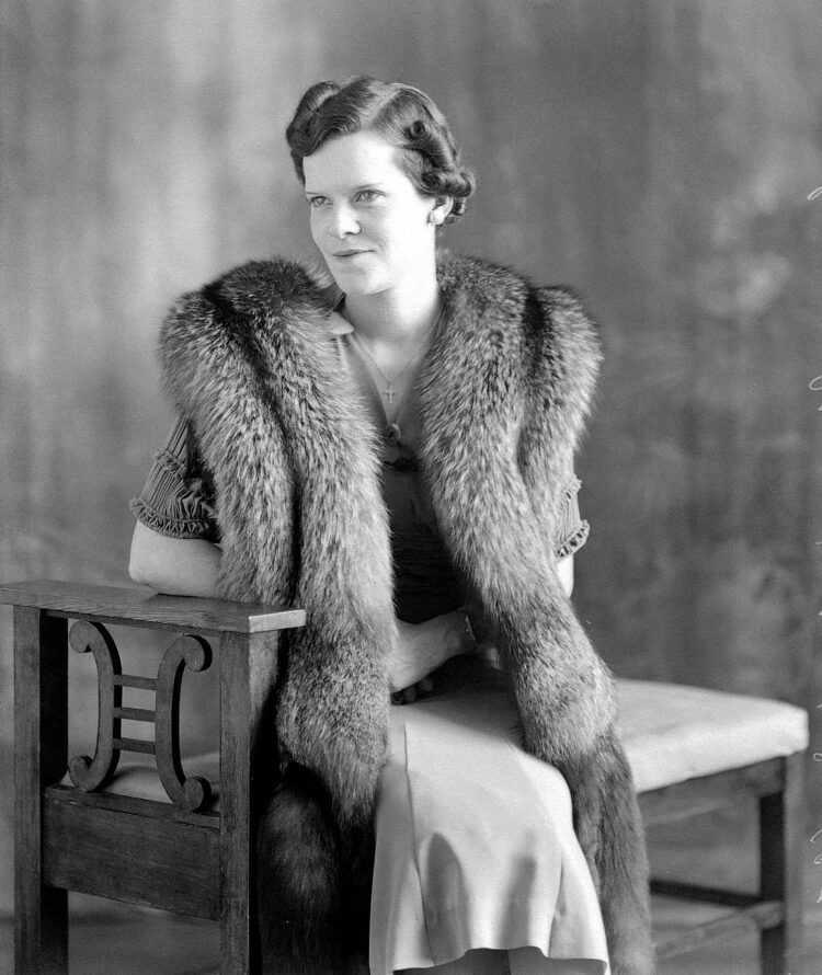 Portrait studio d'une femme assise sur un banc et portant autour de son cou une imposante écharpe de fourrure de renard argenté.
