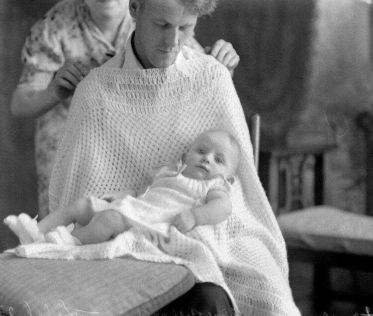 Un homme assis dont le haut du corps et les jambes sont drapés d'un ouvrage brodé tenu par une femme en arrière-plan et sur lequel un bébé est couché.