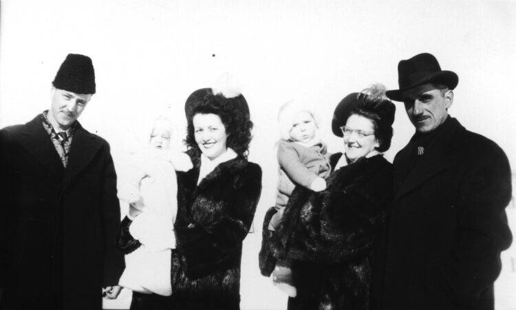 Deux couples vêtus élégamment, avec leur bébé, posant à l'extérieur, en hiver, dans un cliché surexposé.