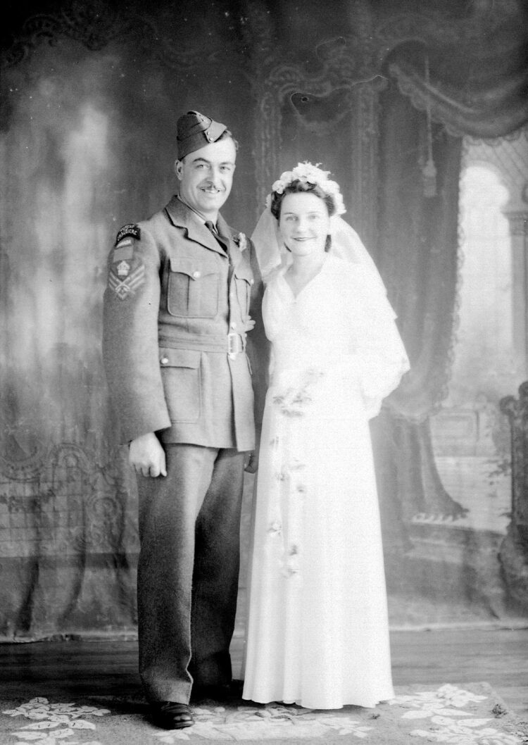 Portrait de studio d'un couple de mariés, la femme portant une robe blanche et l'homme un costume de militaire sur lequel est inscrit "Régiment Chaudière".