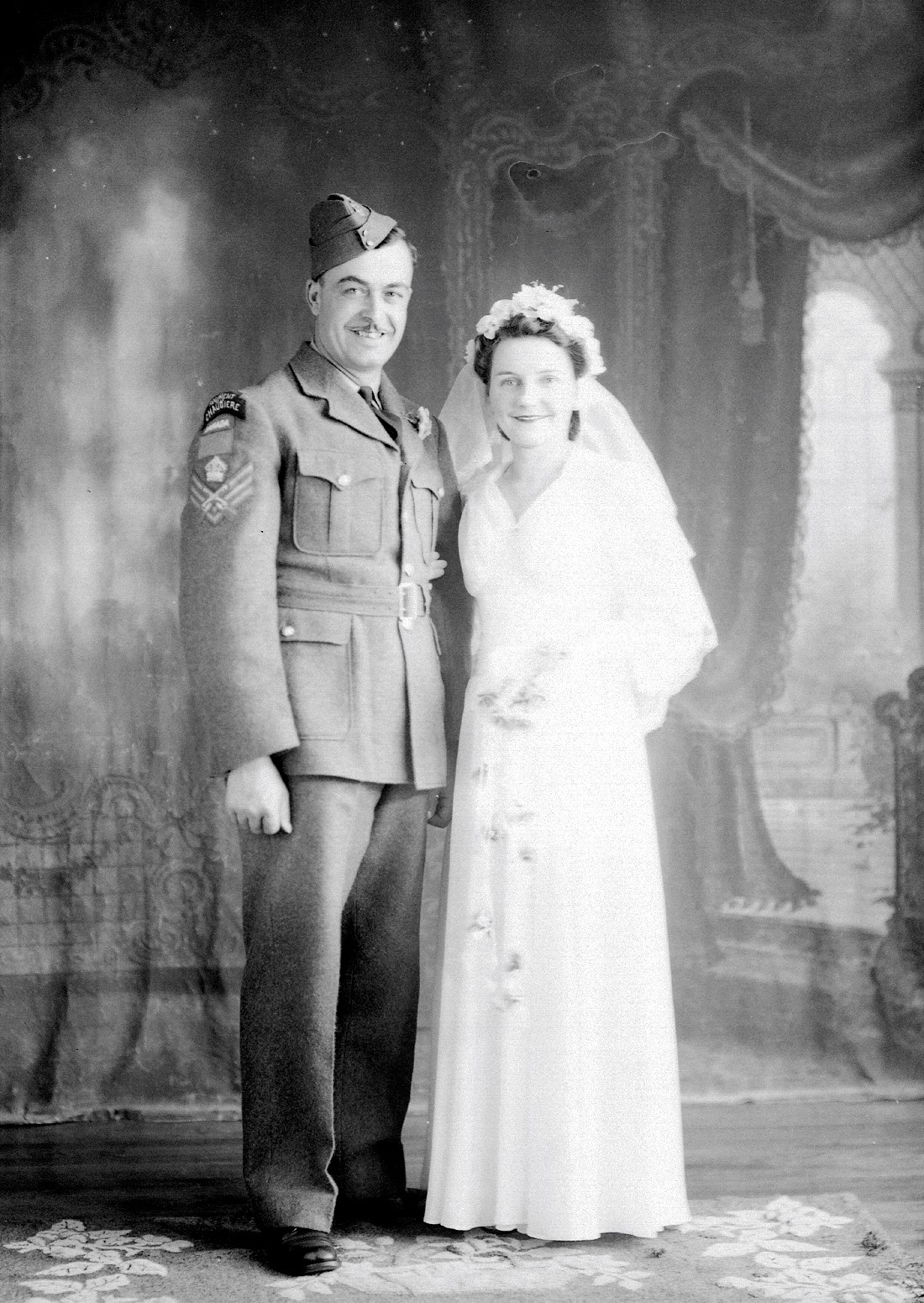 Portrait de studio d'un couple de mariés, la femme portant une robe blanche et l'homme un costume de militaire sur lequel est inscrit "Régiment Chaudière".