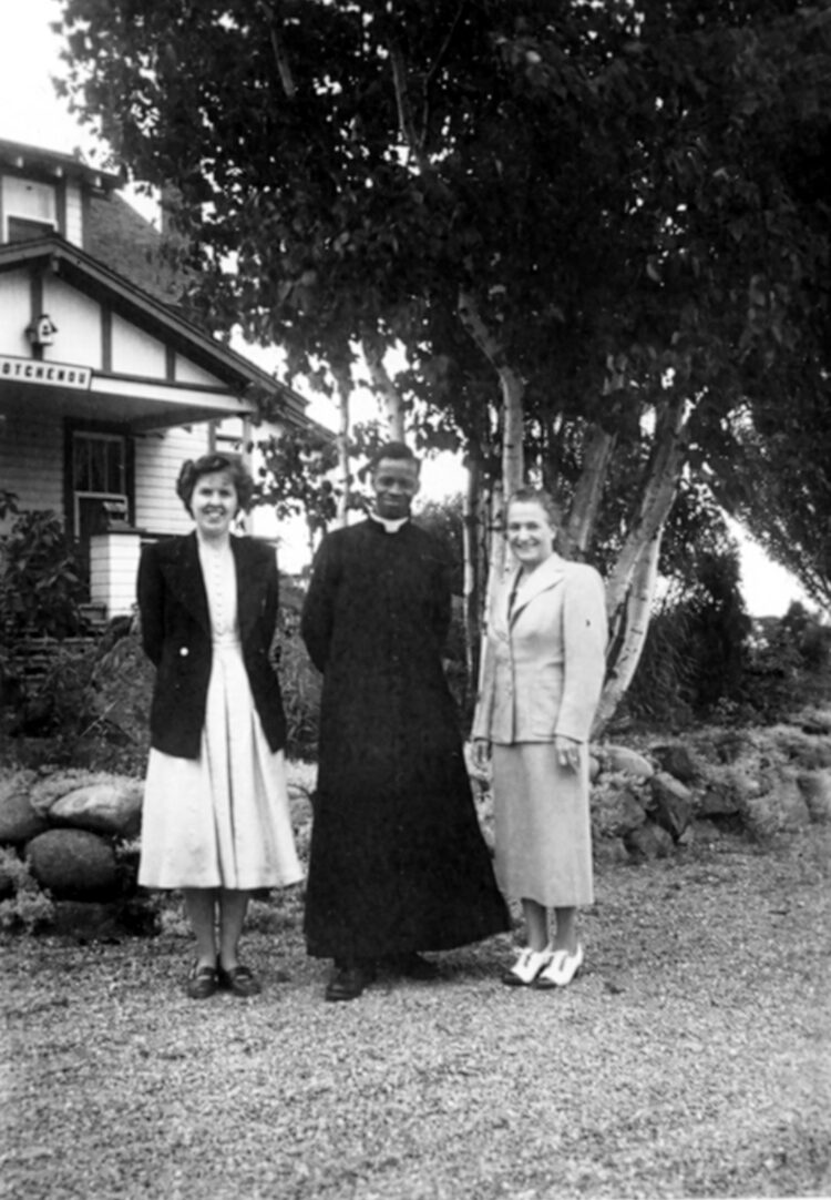 Portrait en extérieur d'un religieux à la peau noire flanqué de deux jeunes femmes blanches.