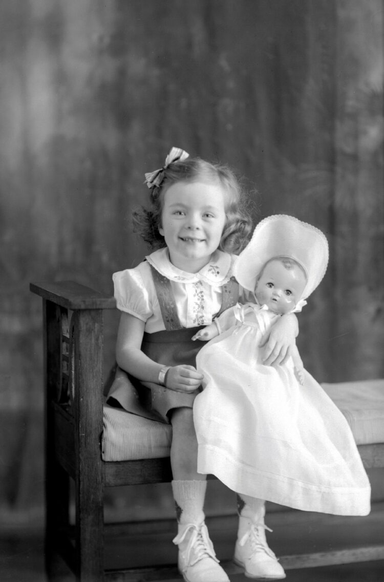Portrait studio d'une fillette assise sur un banc et souriant à la caméra tout en tenant une poupée dans ses bras.