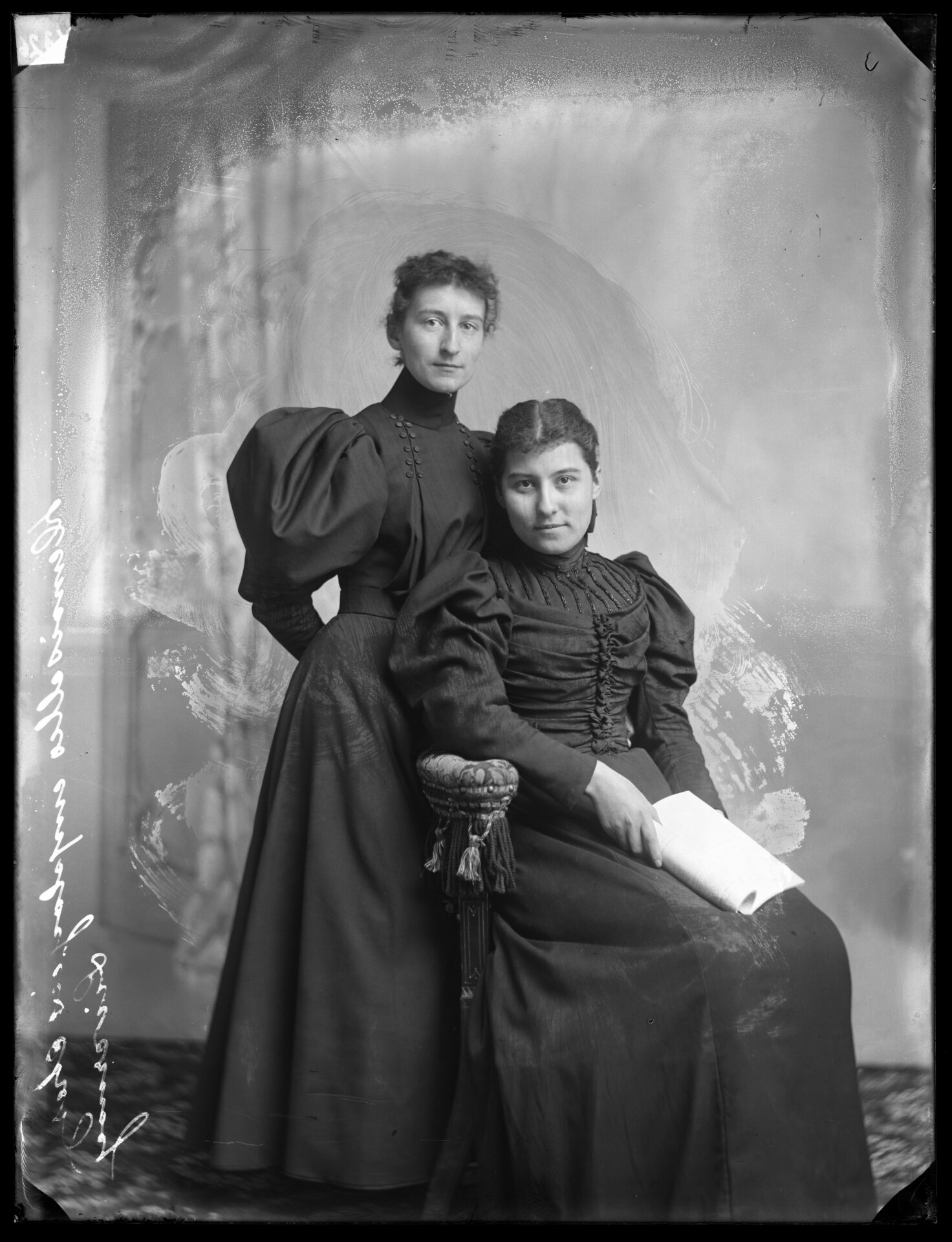 Portrait de deux jeunes femmes en robes foncées en studio dont l'une est debout et l'autre assise devant et tenant un livre dans ses mains.